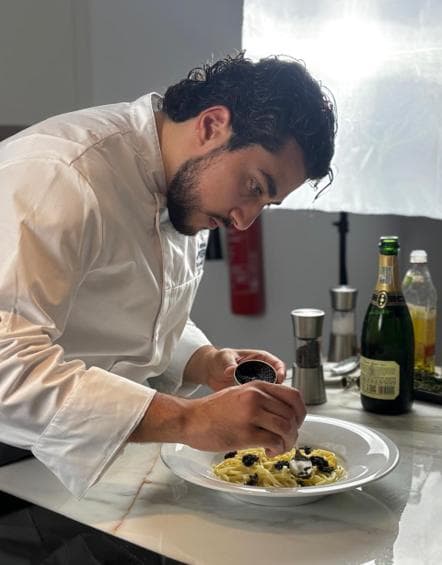A chef plating spaghettini with black caviar beside a bottle of Champagne