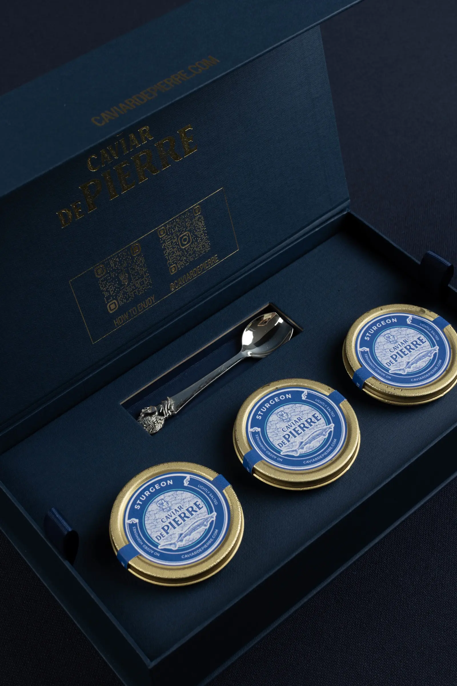An open navy Caviar De Pierre box with the gold logo in its lid, three sturgeon caviar tins and a silver spoon